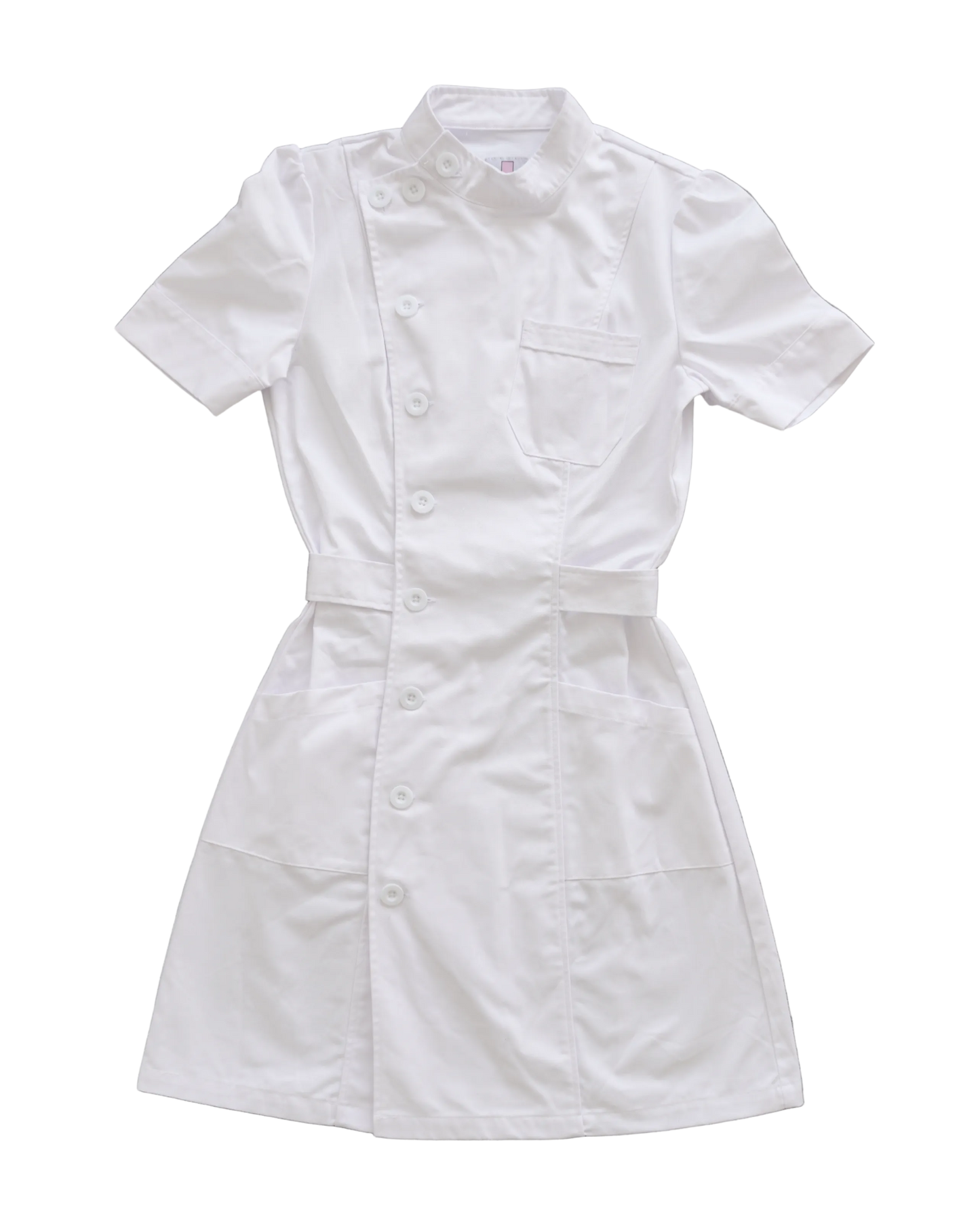 A white short-sleeve nurse scrub dress with a button front, chest pocket, and waist tie, laid flat on a textured gray concrete background.